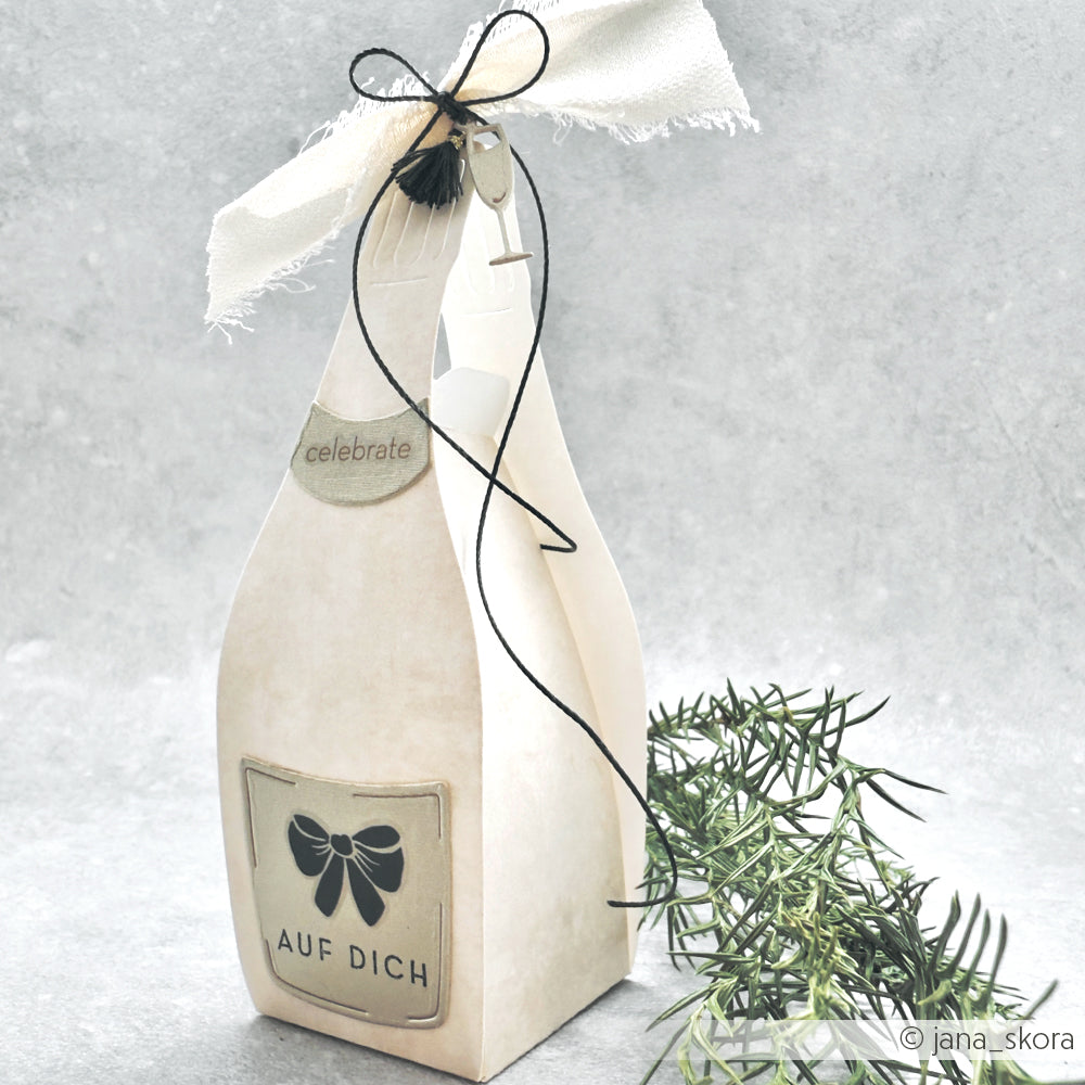 Geschenkbox in Form einer Sektflasche von Alexandra Renke, hergestellt aus Designpapier und veredelt mit der Motivstanze Champagner Box, beschriftet mit "Auf Dich", verziert mit einer Stoffschleife und Grünzeug.