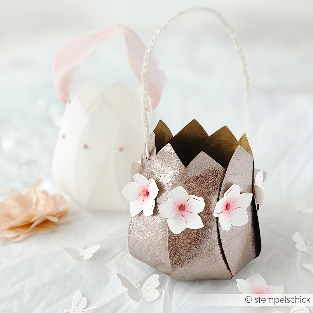 Metallic-pinkes Papierkörbchen aus Designpapier von Alexandra Renke, verziert mit weißen Blumen, mit einem weißen Korb im Hintergrund - ideal für kreative DieCut Systeme oder Motivstanze Blütenkörbchen.