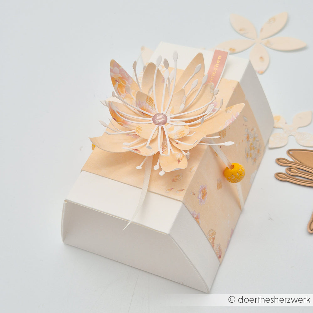 Eine weiße Geschenkbox mit einer blumigen Dekoration in Pfirsichfarben aus Designpapieren von Alexandra Renke, verziert mit einem passenden Band, präsentiert vor einem hellen Hintergrund.
