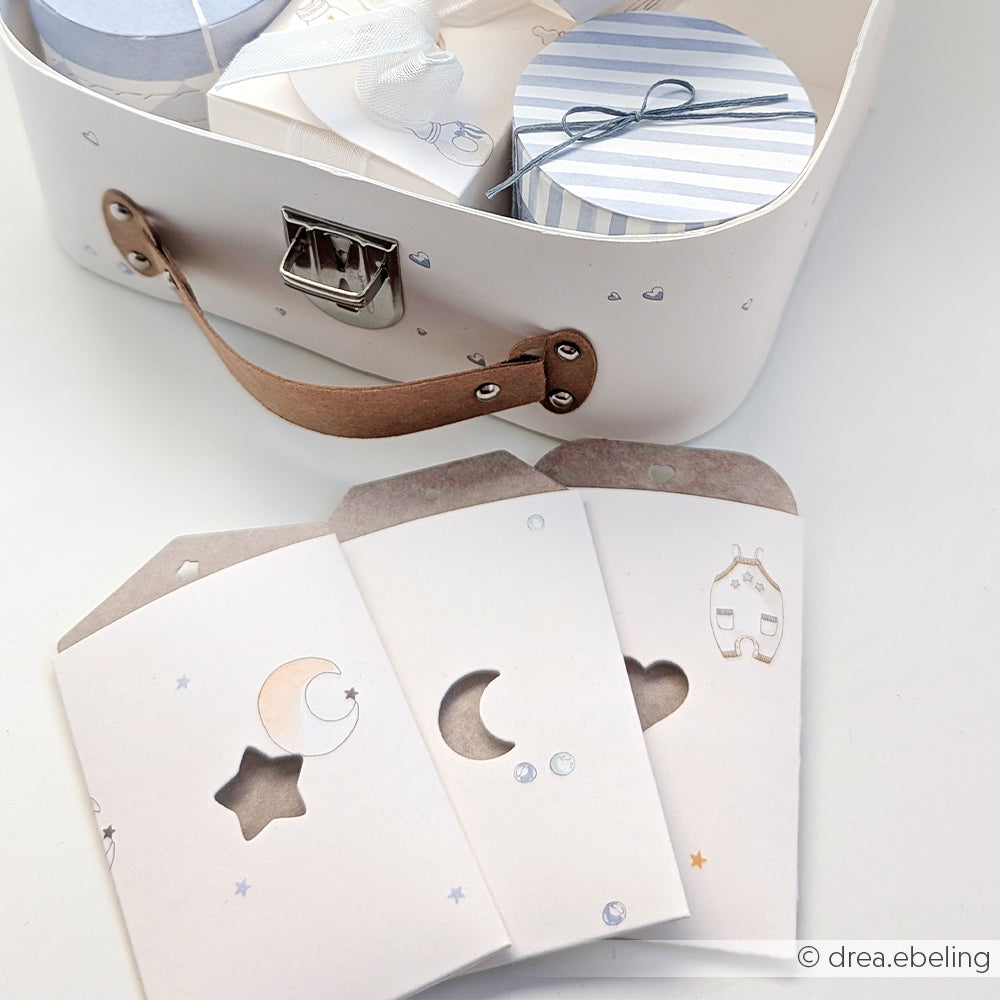 Weißer Koffer von Alexandra Renke mit drei pastellfarbenen Umschlägen mit Stern-, Mond- und Hausausschnitten sowie gestreiften runden Schachteln - perfekt für Ihre Kreativität oder zum Mischen und Kombinieren in Kollektionen oder Bündeln.