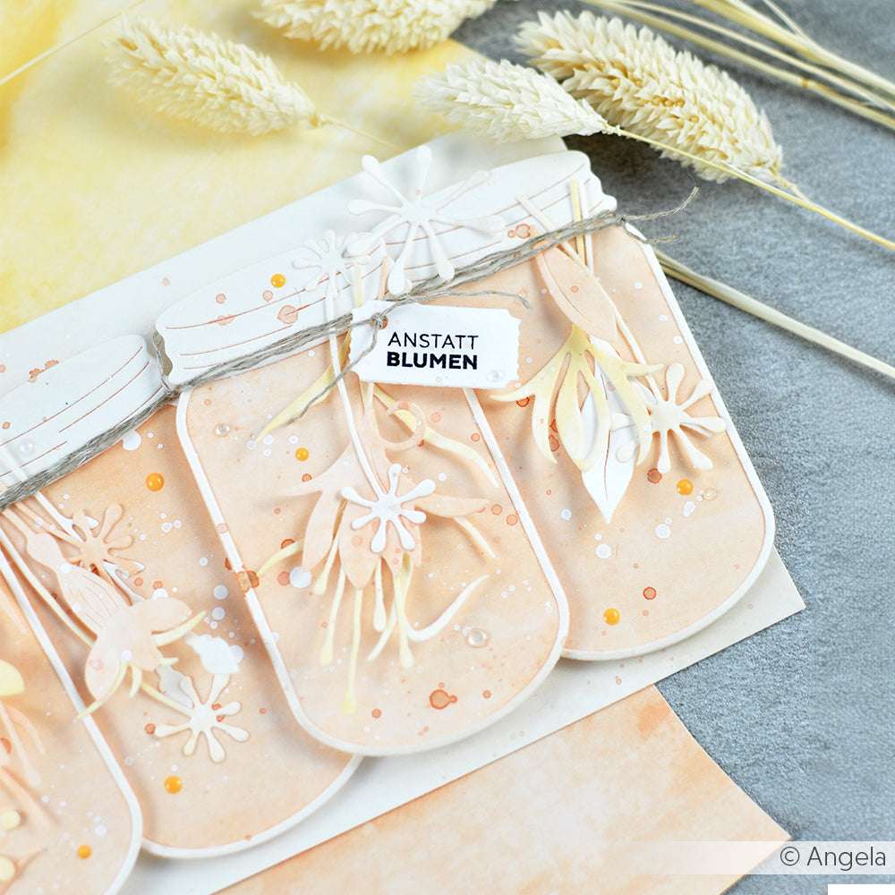 Drei pastellfarbene Papiergefäße aus Designpapier von Alexandra Renke, die mit dem Marmeladengläser-Stanzer mit ausgeschnittenen Blumen verziert, mit "ANSTATT BLUMEN" beschriftet und neben getrockneten Gräsern ausgestellt wurden.