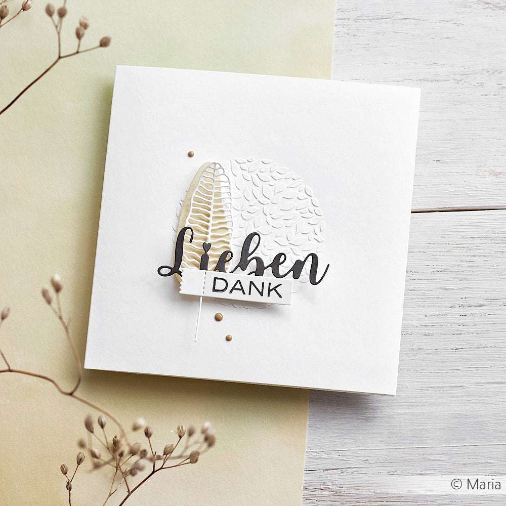 Quadratische Karte von Alexandra Renke, hergestellt mit strukturierten weißen und goldenen Designpapieren. Die Karte trägt den Schriftzug "Lieben DANK" und ist auf einem hellen Holzhintergrund angebracht.