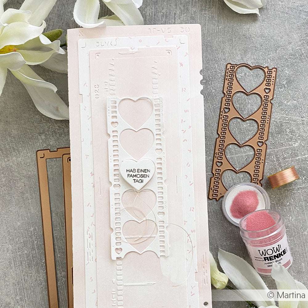 Eine handgefertigte Karte mit einer Filmstreifen-Stanzung aus der Motivstanze Film Herzen von Alexandra Renke, akzentuiert durch rosa Prägepulver und weiße Blumen im Hintergrund.