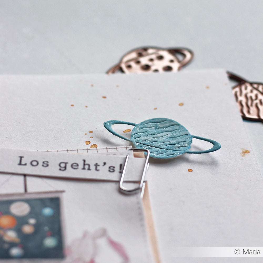 Papier mit einer blauen Planetenklammer, die mit Alexandra Renke Motivstanze 3 Planeten hergestellt wurde, auf der neben Zeichnungen zum Thema Weltraum auch "Los gehts!" steht.
