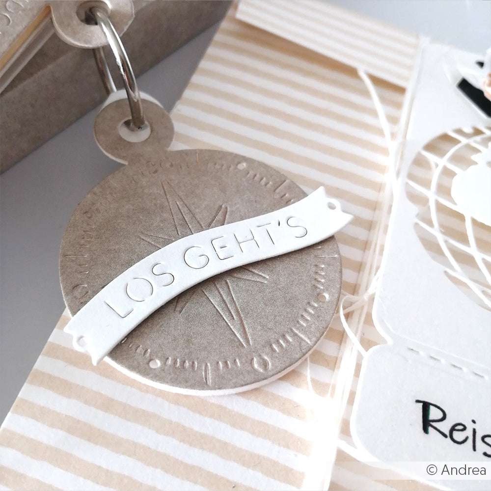 Runder beiger Anhänger von Alexandra Renke mit "LOS GEHTS" auf weißem Banner und gestreiftem Hintergrund, befestigt an einem Metallring - gebastelt aus Designpapieren und akzentuiert mit der Motivstanze Abhefter Kompass DieCut Systeme.