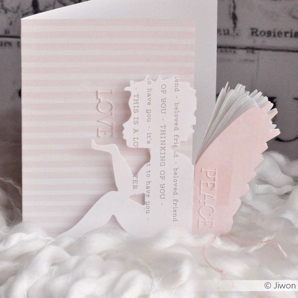 Eine engelsförmige Karte mit Alexandra Renke Motivstanze Engelsflügel und Designpapieren, mit LOVE und PEACE Text, ist auf einem weichen weißen strukturierten Hintergrund abgebildet.