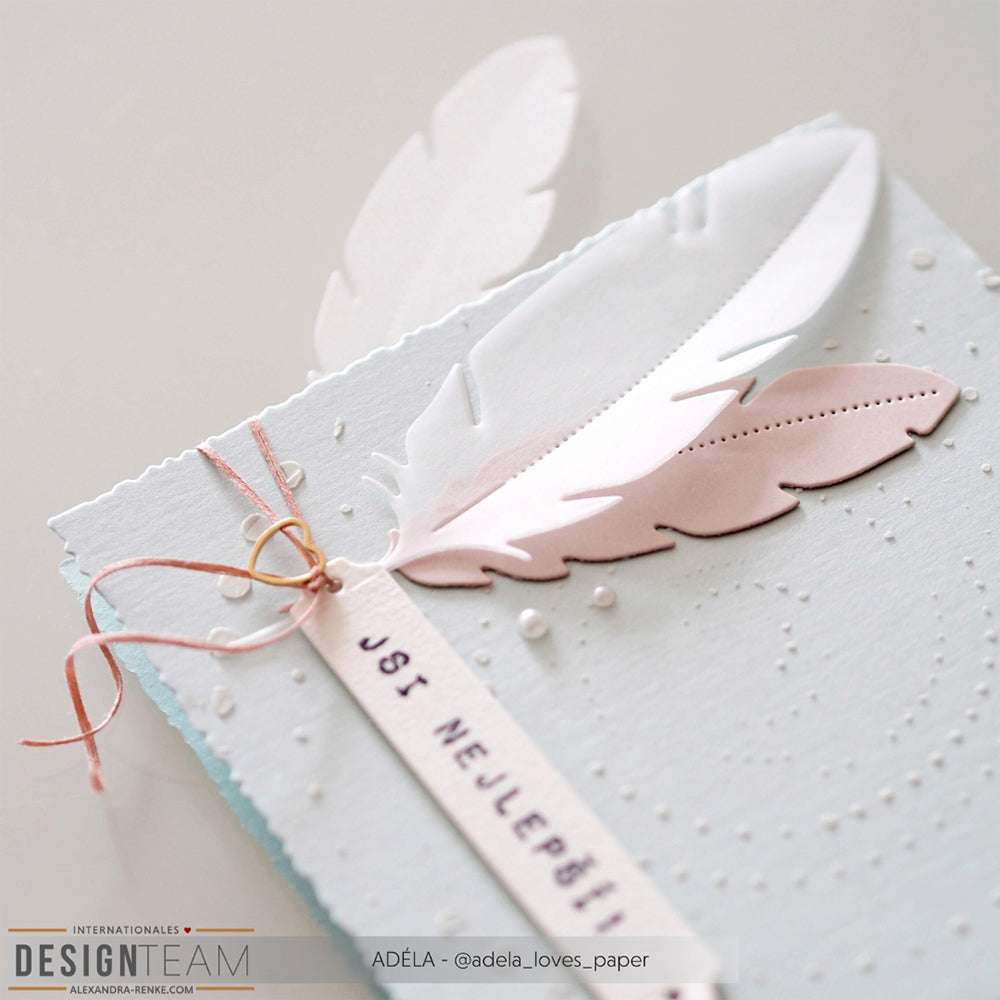 Handgefertigte Karte mit weißen und rosafarbenen Papierfedern, die mit der Alexandra Renke Motivstanze Feder hergestellt wurden, genähten Details und einem mit Schnur gebundenen Anhänger "JOS NEJLEPŠÍ".