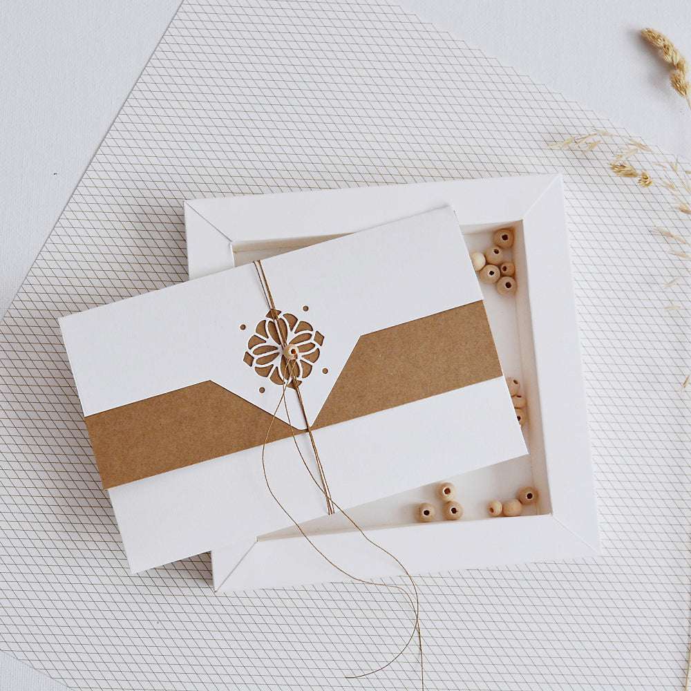 Weiße Einladungskarte von Alexandra Renke mit brauner Banderole und eleganter Motivstanze, die auf einem weißen Tablett mit kleinen Holzperlen steht.