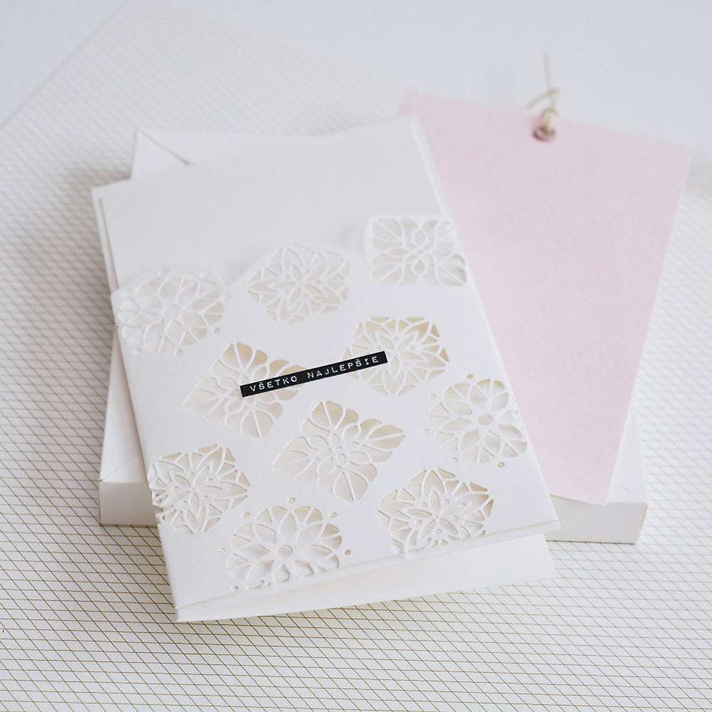 Weiße Grußkarte von Alexandra Renke, hergestellt aus Designpapieren mit detaillierten Motivstanze Ornament mit Kontur Ausschnitten und einem schwarzen Etikett, neben einem blassrosa Notizblock angezeigt.