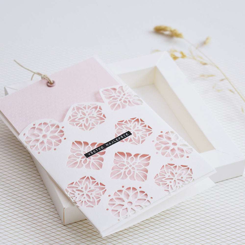Eine zarte weiße Karte mit rosafarbenen Akzenten und gestanzten Blumenmustern von Alexandra Renke, präsentiert auf einer offenen weißen Schachtel.