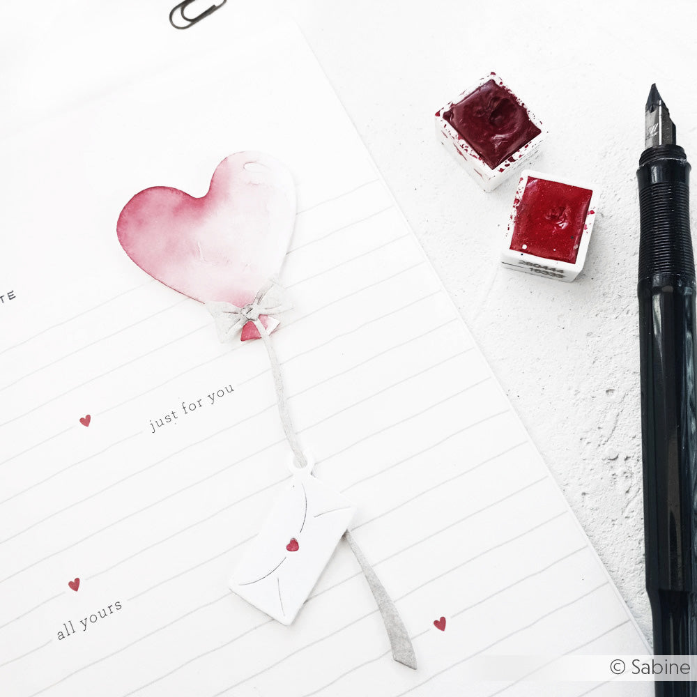Eine Notizbuchseite mit einem aquarellierten Herz, einem Umschlag, einem Stift und roter Farbe im Stil eines Liebesbriefs - perfekt, um mit der Motivstanze Herzballon Grüße von Alexandra Renke kreative Akzente zu setzen.