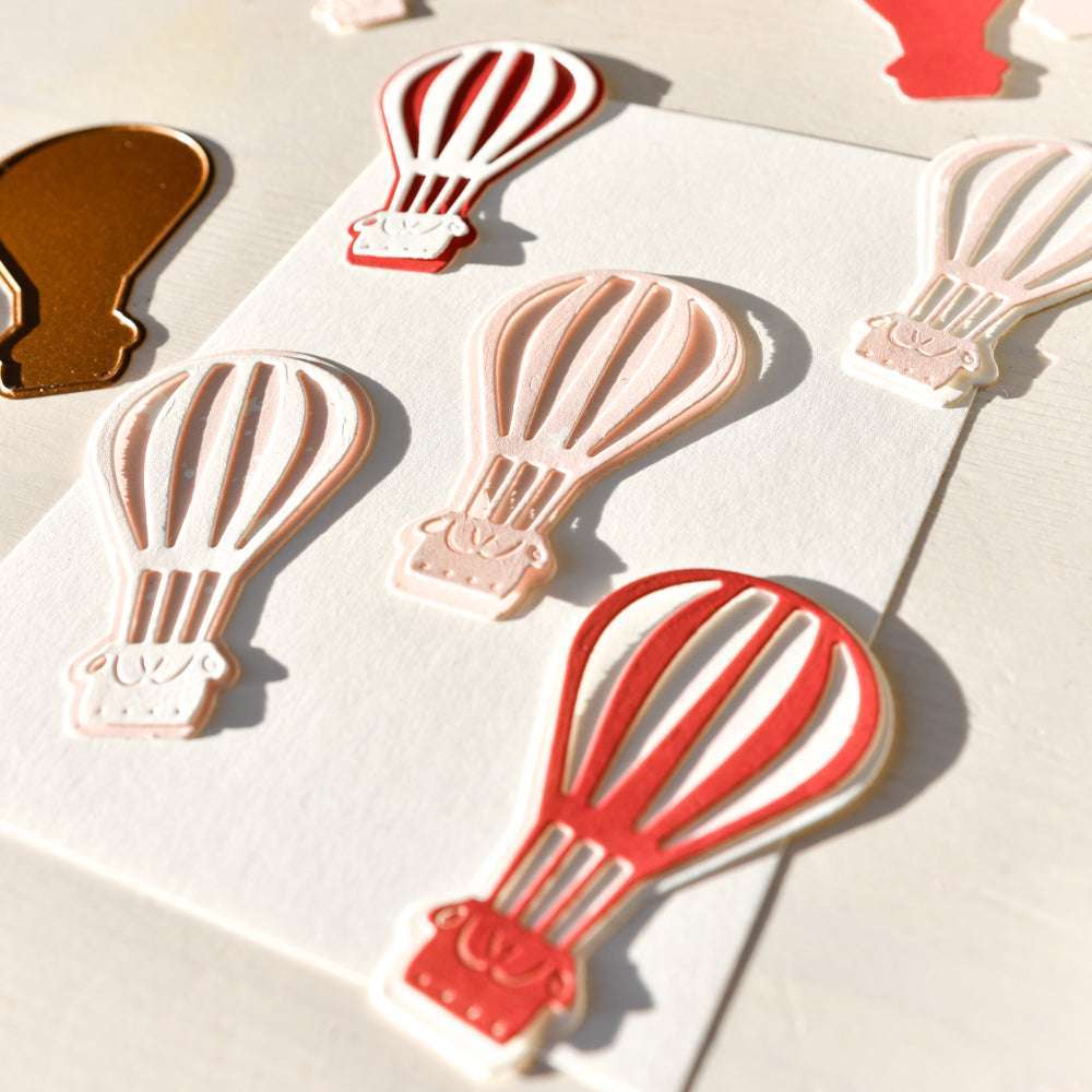 Die mit der Alexandra Renke Motivstanze Heißluftballon aus schimmernden roten, rosa und goldenen Designpapieren gestalteten Papierausschnitte von Heißluftballons ruhen auf einer weißen Fläche im hellen Sonnenlicht.