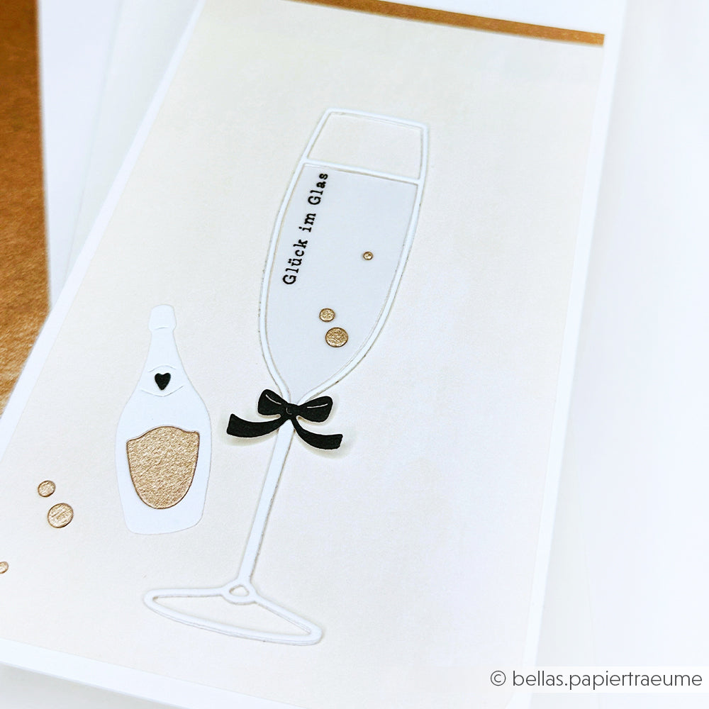 Minimalistische Karte mit einem Sektglas, einer kleinen Flasche, goldenen Akzenten und dem Text "Glück im Glas" - perfekt, um Ihre Alexandra Renke Motivstanze Fröhliche Drinks oder DieCut Systeme auf Designpapier zu präsentieren.