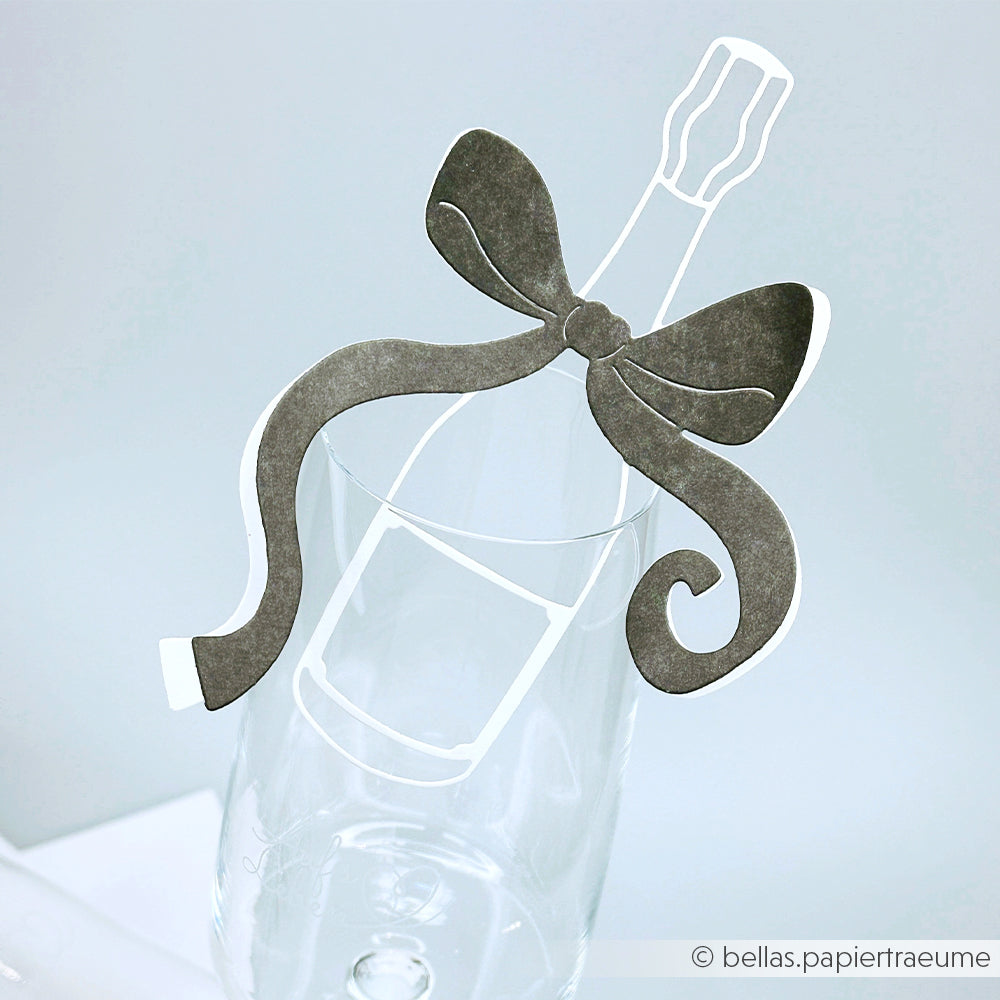 Ein mit einer Papierschleife verziertes Sektglas, das von Alexandra Renke Designpapiere mit der Motivstanze Große Champagnerflasche gefertigt und am Rand befestigt wurde.