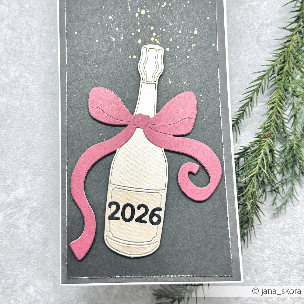 Eine Papiersektflasche aus Alexandra Renke Designpapieren mit rosa Schleife und 2026-Etikett, gestaltet mit der Motivstanze Große Champagnerflasche, steht auf einem dunklen Hintergrund mit Tannenzweigen.