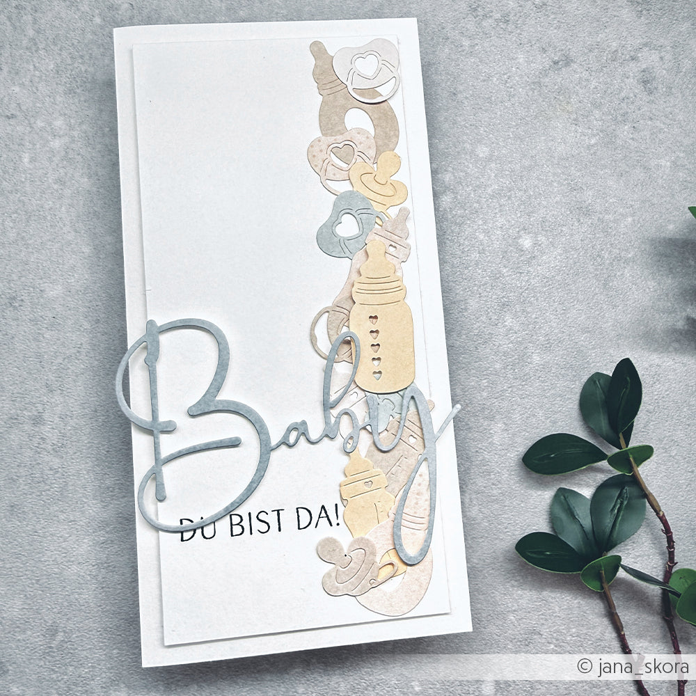 Handgefertigte Karte mit pastellfarbenen Babyartikeln, dem Text "Baby" und einem Zweig mit grünen Blättern auf grauem Grund, hergestellt mit Alexandra Renke Designpapieren und dem Motivstanze Fläschen Set für zusätzlichen Charme.