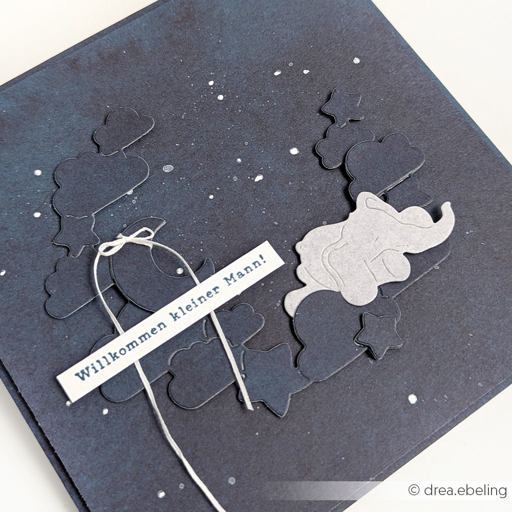 Die dunkelblaue Karte von Alexandra Renke zeigt Wolken, Sterne, einen grauen Elefanten und "Willkommen kleiner Mann!" auf einem weißen Etikett - gefertigt mit hochwertigen Designpapieren und der Motivstanze Nachthimmelkranz für charmante Details.