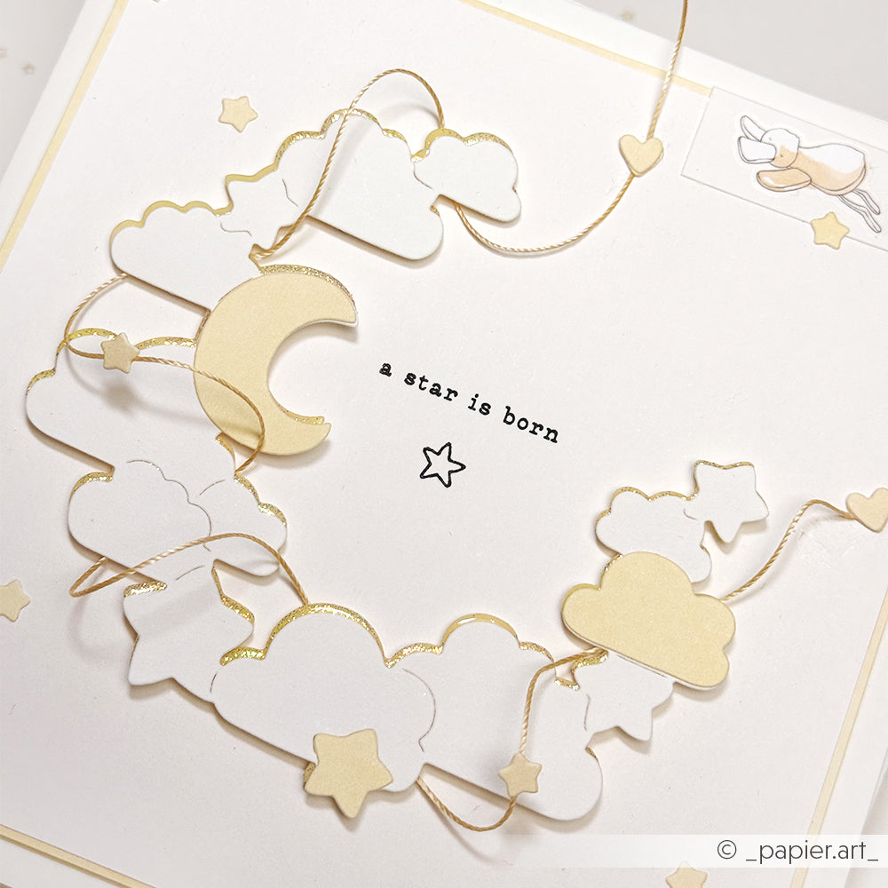 Handgemachte Karte von Alexandra Renke aus Designpapier mit Wolken, Sternen und einer Mondsichel. In der Mitte steht "Ein Stern ist geboren". Dekorative Details mit der Motivstanze Nachthimmelkranz sorgen für himmlischen Charme.
