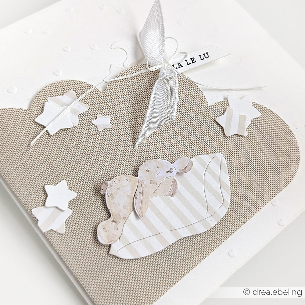 Eine Babykarte von Alexandra Renke mit einer Stoffwolke aus der Motivstanze Kartenaufleger Wolke, Papiersternen und einem Häschen, das sich an eine gestreifte Decke kuschelt, verziert mit einem weißen Band.