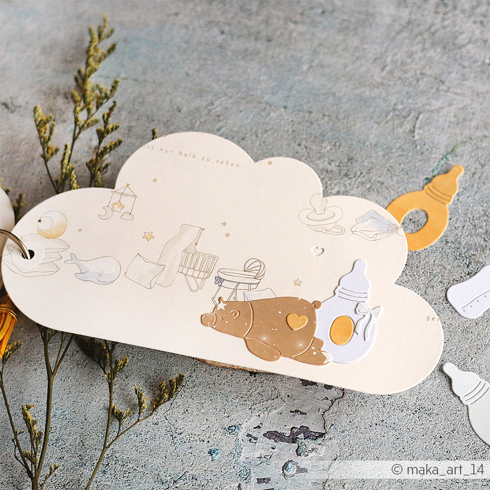 Eine wolkenförmige Karte, die mit der Alexandra Renke Motivstanze Wolke hergestellt wurde, zeigt Babyartikel, einen Bären, eine Flasche und subtile botanische Akzente auf einer grauen Oberfläche.