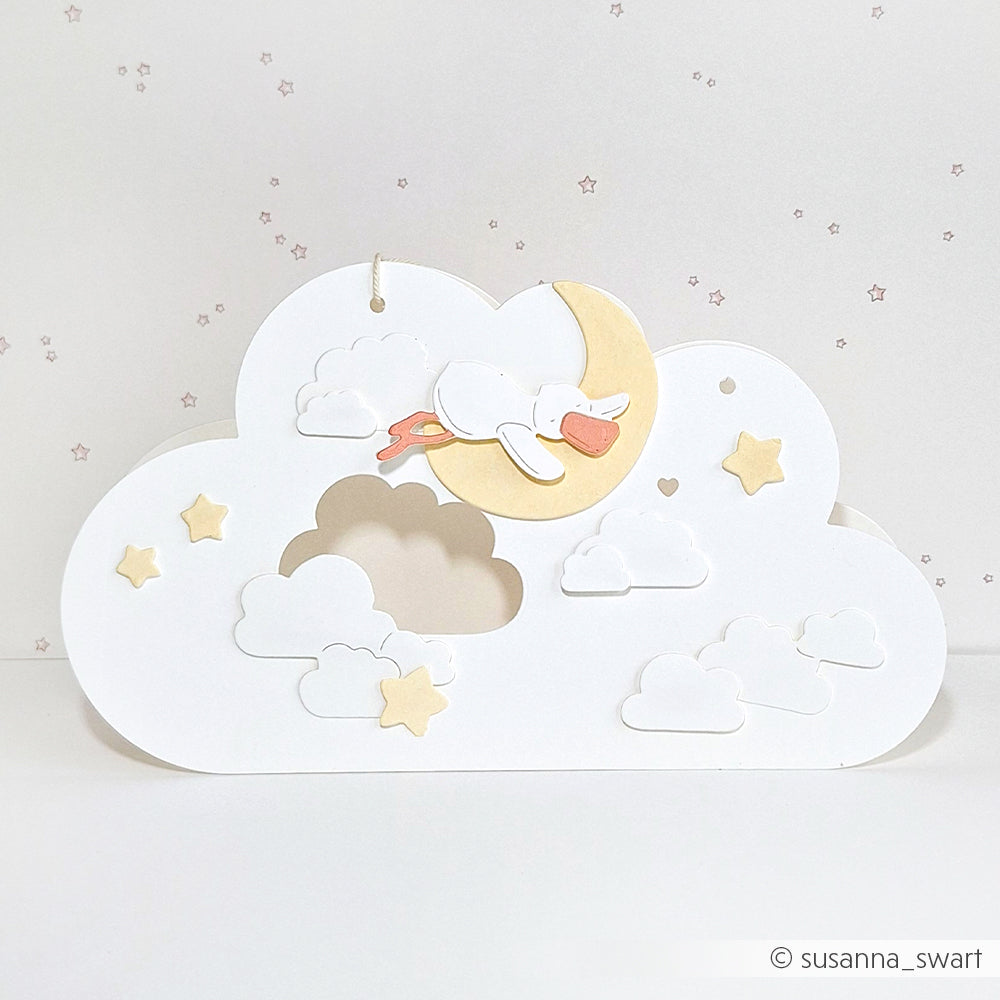 Scherenschnitt eines Storches auf einem Mond, umgeben von Wolken und Sternen, hergestellt mit Alexandra Renke Designpapiere, vor einem hellen Hintergrund.