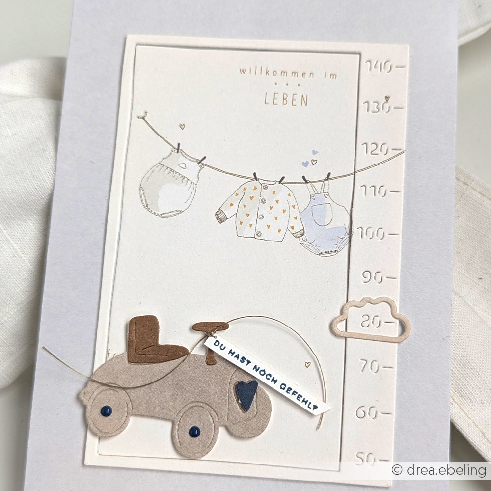 Handgefertigte Babykarte von Alexandra Renke mit gestanzten Kleidungsstücken an einer Leine, einem niedlichen Spielzeugauto und einem deutschen Willkommensgruß für Neugeborene - gefertigt mit Motivstanze-Details für eine ganz besondere Note.