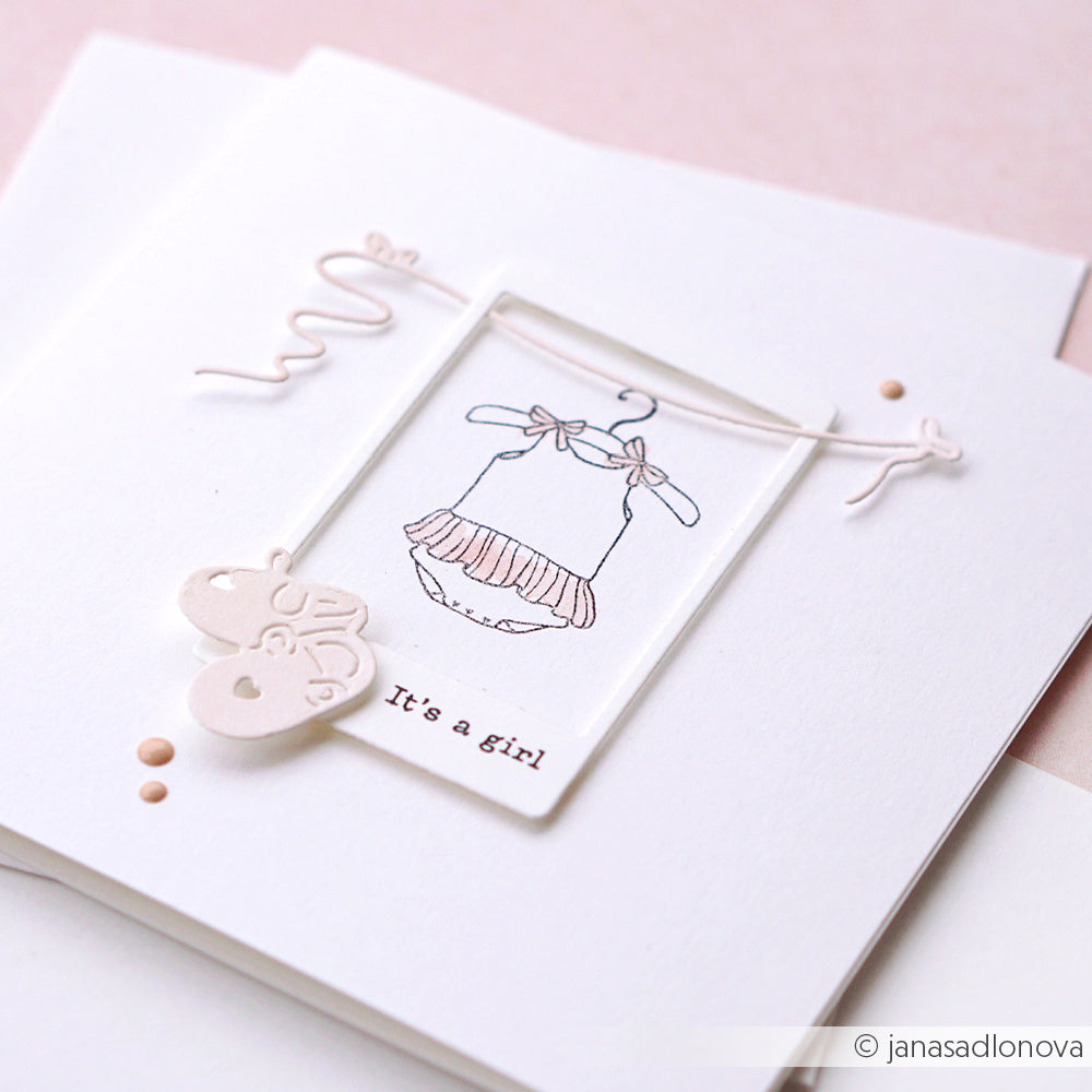 Minimalistische Karte von Alexandra Renke mit einer Skizze eines Babykleides, zwei Herzanhängern und dem Text "It's a girl" auf hochwertigen Designpapieren, akzentuiert mit einer Motivstanze Kleine Schuhe für zusätzlichen Charme.