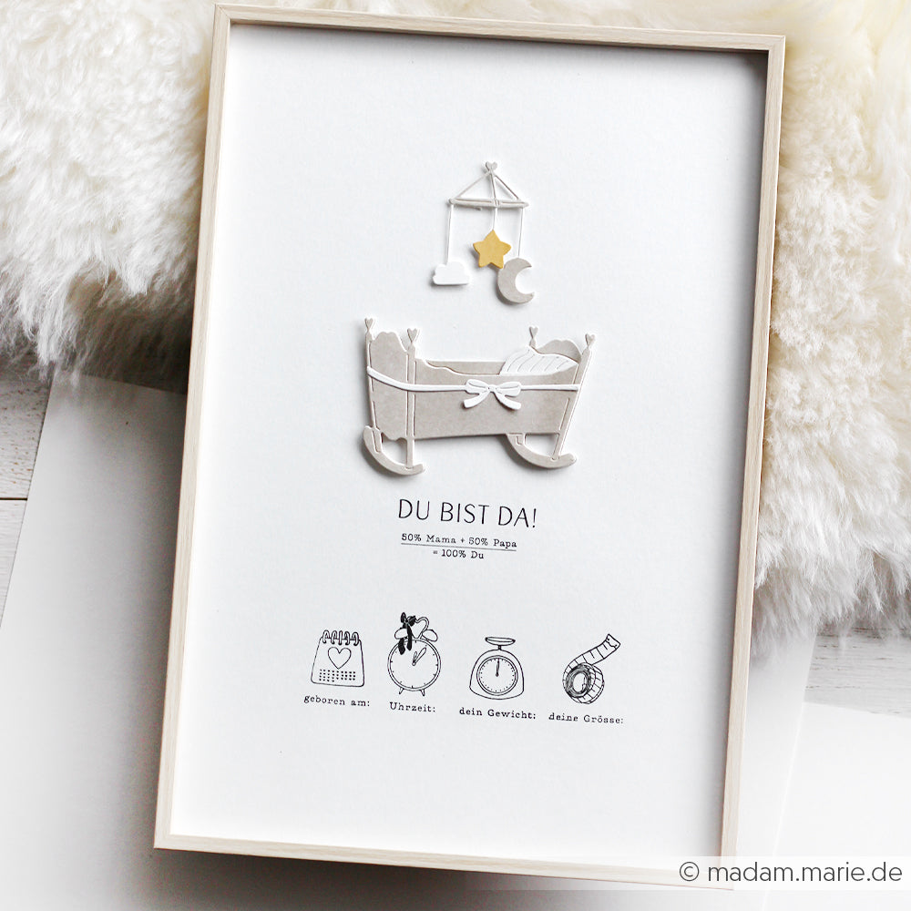 Gerahmter Druck mit einer 3D-Wiege, Sternen und Symbolen zur Geburt, beschriftet mit "Du bist da!" von Alexandra Renke mit Designpapieren und der Motivstanze Herzliche Wiege für ein charmantes Finish.