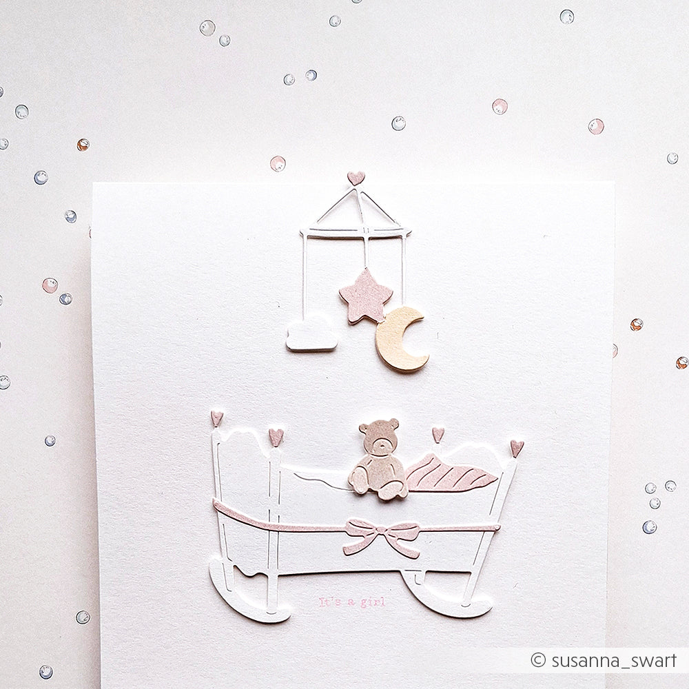 Minimalistische Karte für ein kleines Mädchen von Alexandra Renke, gestaltet mit Designpapieren und verziert mit einem Kinderbett, einem Teddybär, einem Mobile und zartrosa Akzenten auf einem weiß gepunkteten Hintergrund. Veredelt mit Dies Stanzen für einen eleganten, zarten Touch.