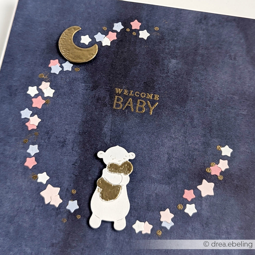 Eine Karte mit "Welcome Baby", einem Bären, der ein Herz umarmt, einem goldenen Mond und pastellfarbenen Sternen auf dunklem Hintergrund, gestaltet mit Alexandra Renke Designpapieren für zusätzlichen Charme.