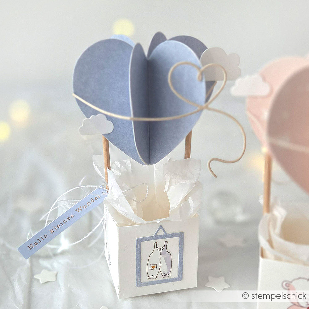 Ein blauer, herzförmiger Heißluftballon aus Papier von Alexandra Renke, hergestellt mit Motivstanze, präsentiert in einer weißen Schachtel mit Wolken und einem Baby-Anhänger.