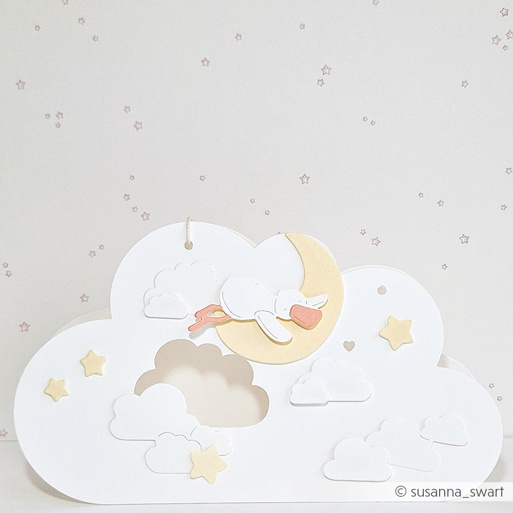 Scherenschnitt eines schlafenden Storches auf einem Mond mit Wolken und Sternen auf pastellfarbenem Hintergrund - hergestellt mit der Alexandra Renke Motivstanze für präzise Formen auf hochwertigem Designpapier.