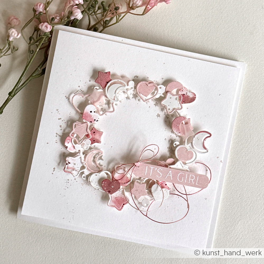 Die quadratische Karte von Alexandra Renke zeigt einen rosafarbenen und weißen Kranz aus Sternen, Monden und Herzen mit DieCut Systemen; das Etikett lautet ITS A GIRL. Abgeschlossen mit charmanten Blumen an der Seite.
