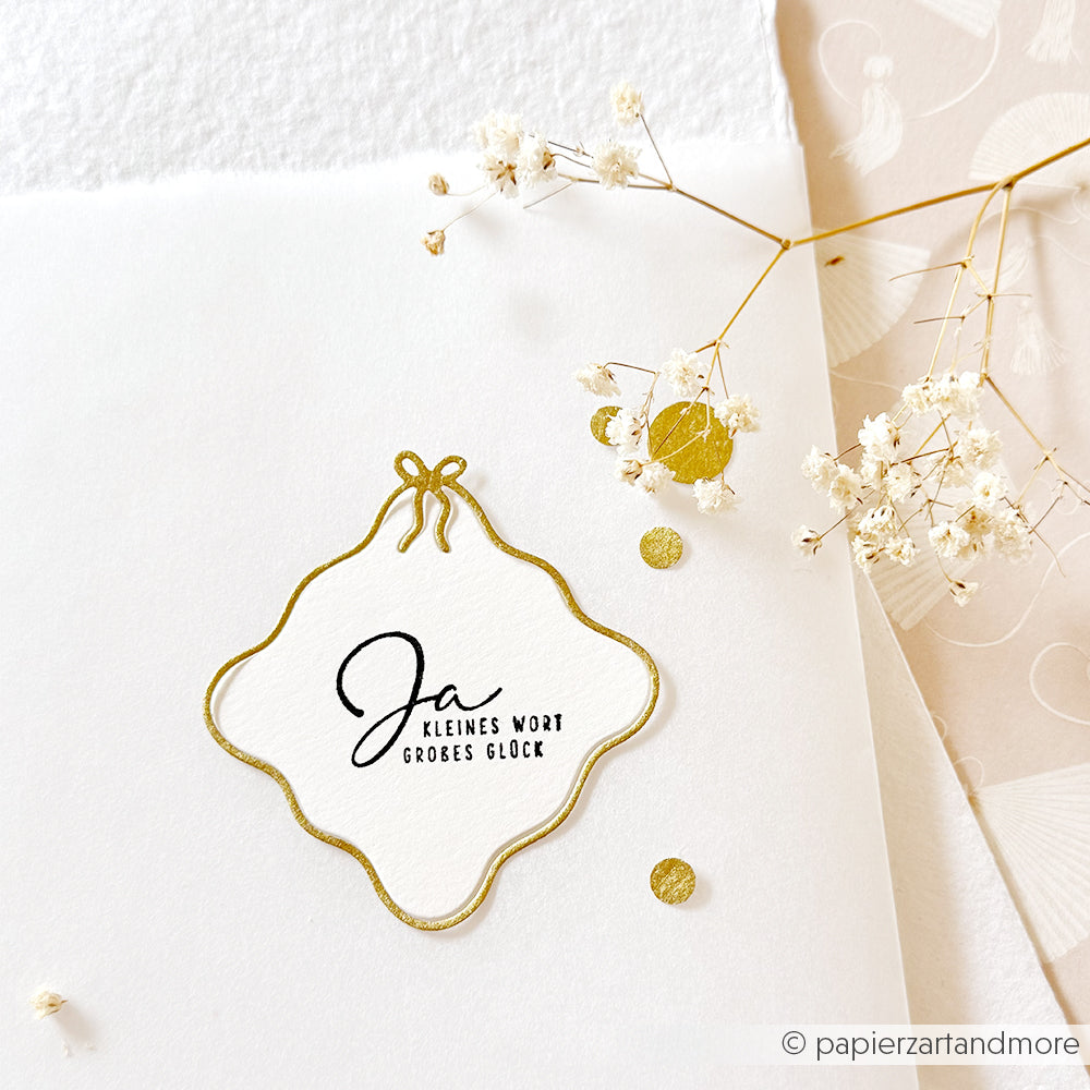 Weiße Karte von Alexandra Renke mit goldenen Akzenten, getrockneten Blumen und dem Text "Ja kleines Wort großes Glück" in eleganter Schrift, verziert mit einer Motivstanze Wellenrand-Rahmen. Ideal für DieCut Systeme oder Stanzen für Designpapier.