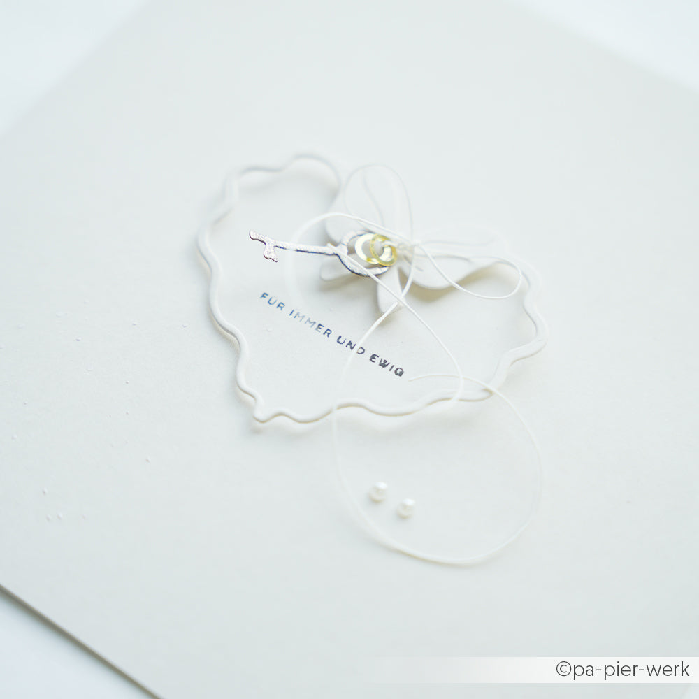 Eine zarte weiße Karte von Alexandra Renke zeigt einen Schlüssel, eine Schleife, ein florales Detail und den Text FÜR IMMER UND EWIG, elegant akzentuiert mit einer Stanzschablone Herzrahmen.