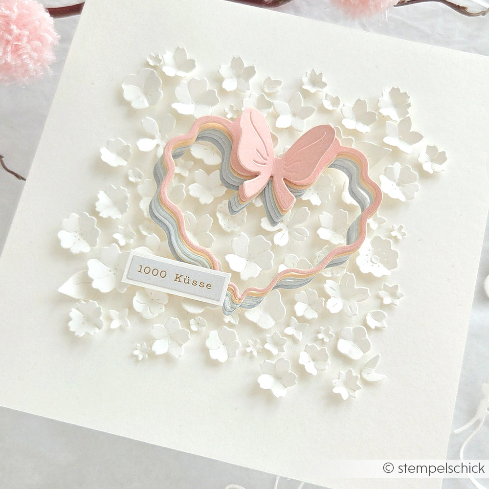 Handgefertigte Karte von Alexandra Renke mit pastellfarbenem Herz und Schleife, hergestellt mit Motivstanze Herz-Rahmen und 200g Premium Designpapieren, verziert mit kleinen weißen Blumen und einem "1000 Küsse"-Anhänger.