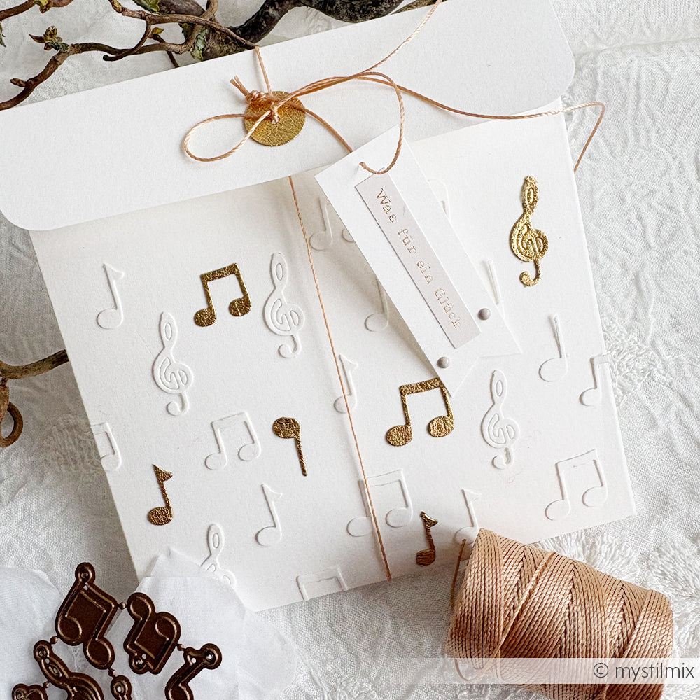 Weiße Karte mit geprägten und goldenen Musiknoten, hergestellt mit Alexandra Renke Motivstanze Noten Anhänger DieCut Systeme, mit einer goldenen Schnur und einem Geschenkanhänger auf einer strukturierten weißen Oberfläche.