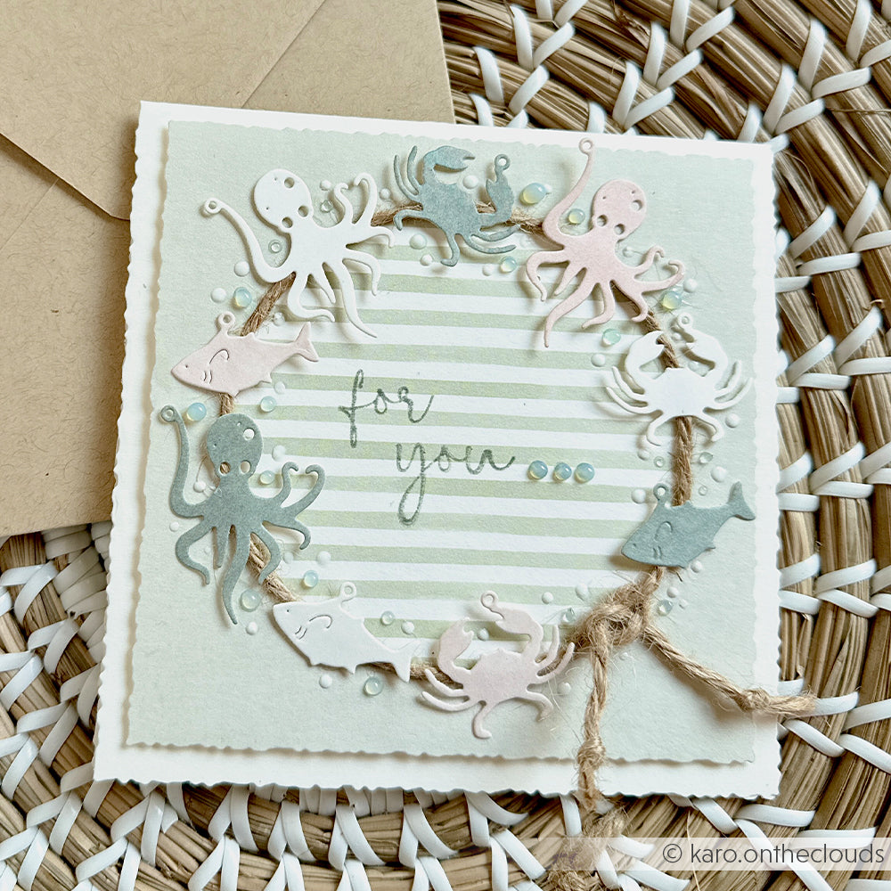 Handgefertigte Karte von Alexandra Renke mit Meeresbewohnern und einem Kranz aus Schnur, verziert mit dem Motivstanze-Anhänger „Meeresspaß“ und eleganten Designpapieren. Die Karte zeigt den Text „for you...“ auf einem gestreiften Hintergrund und wurde auf einer gewebten Unterlage fotografiert.