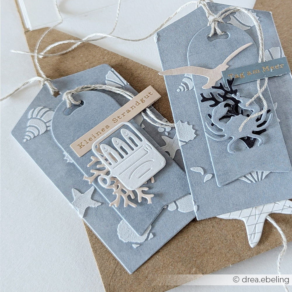 Zwei blaue, handgefertigte Geschenkanhänger von Alexandra Renke, die mit der Motivstanze „Maritime Anhänger“ und Designpapieren gestaltet wurden, sind mit Ausschnitten zum Thema Meer, Schnur und deutschen Etiketten verziert und auf einem braunen Umschlag befestigt.