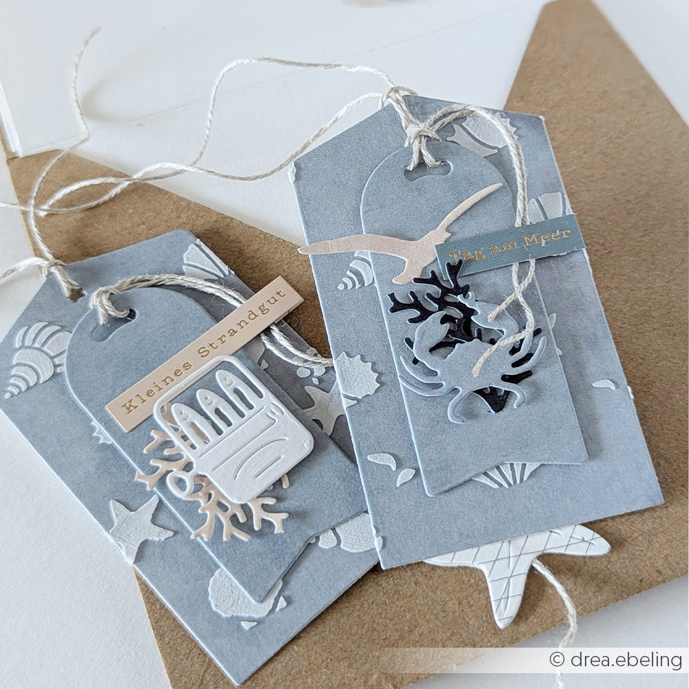 Zwei graue Geschenkanhänger mit Papierausschnitten zum Thema Strand, die mit der Motivstanze „Strandgefühle“ von Alexandra Renke und Designpapier gestaltet wurden, mit Schnur zusammengebunden und an einem braunen Umschlag befestigt.