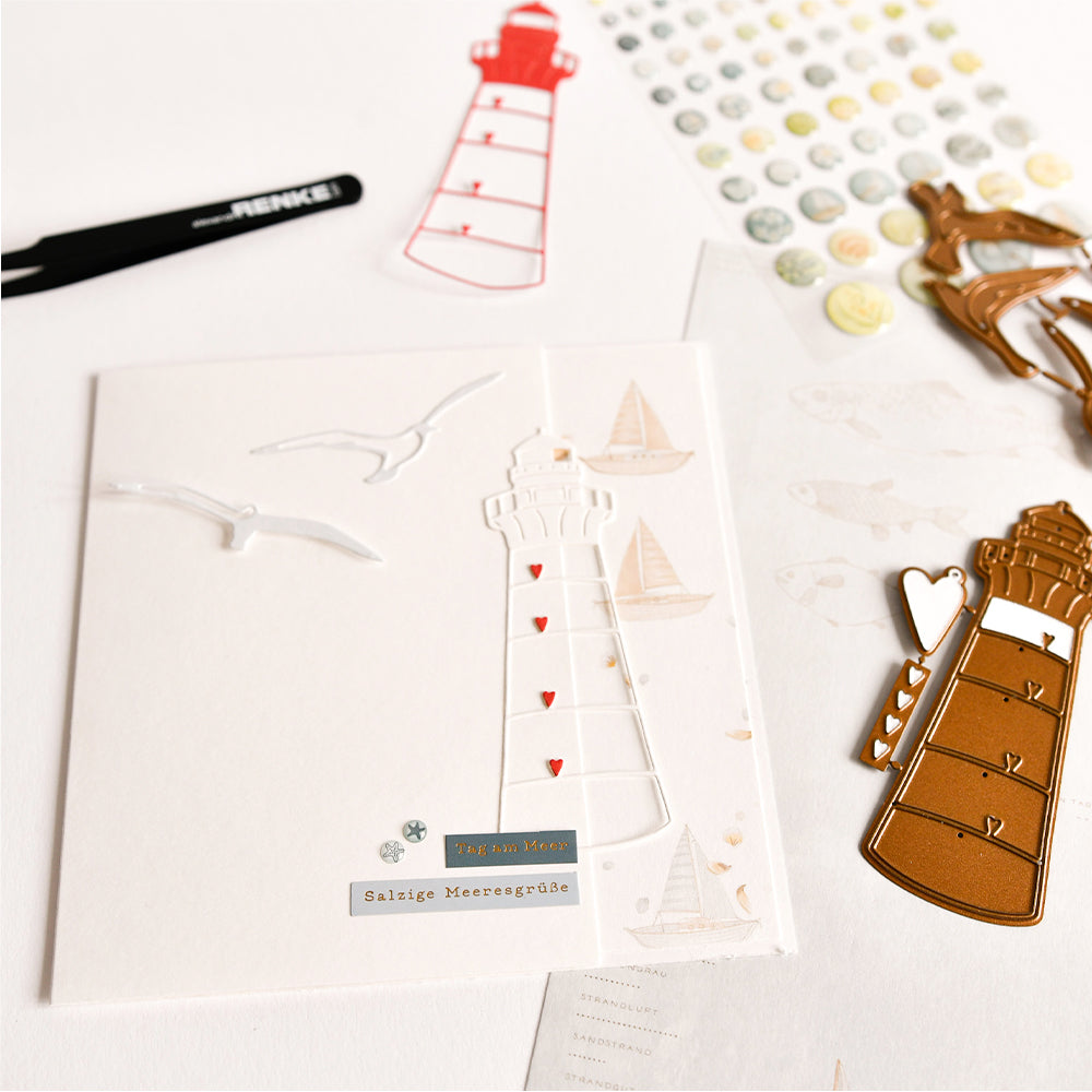 Eine handgefertigte Karte mit einem Leuchtturm, Möwen und maritimen Aufklebern, gestaltet mit Designpapier von Alexandra Renke und der Stanze „Herzlicher Leuchtturm“, präsentiert inmitten verschiedener Bastelwerkzeuge und -materialien.