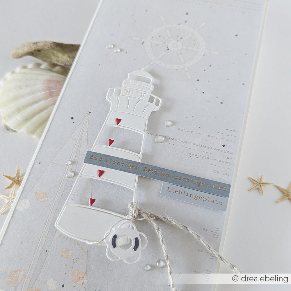 Weiße Karte aus hochwertigen Designpapieren von Alexandra Renke, verziert mit einem Leuchtturmmotiv, das mit der Motivstanze „Herzlicher Leuchtturm“ gestaltet wurde. Akzentuiert mit kleinen roten Herzchen und deutschen Textetiketten, abgerundet durch eine Muschel und maritime Dekorelemente für einen bezaubernden maritimen Touch.
