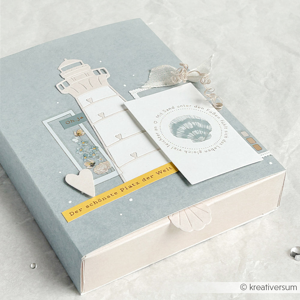 Hellblaue Geschenkbox mit dem Motivstanze-Motiv „Herzlicher Leuchtturm“ von Alexandra Renke, Herzmotiv und Anhänger. Ideal für Bastelfreunde, die Stanzsysteme oder Stanzformen für Designpapier verwenden.