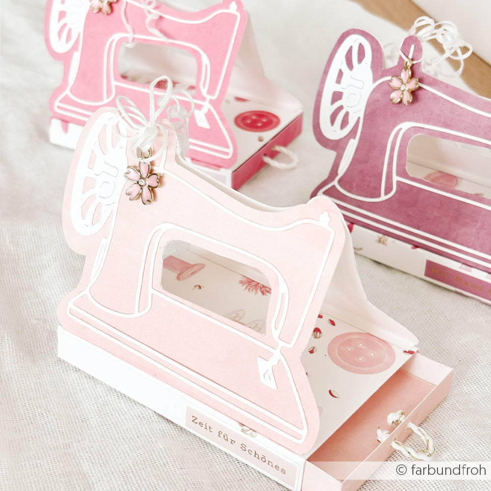 Rosa und lila Geschenkboxen in Form einer Nähmaschine mit Blumenanhängern, hergestellt aus Designpapieren von Alexandra Renke und mit der Motivstanze „Nähmaschine“ von DieCut Systeme für dekorative Details.