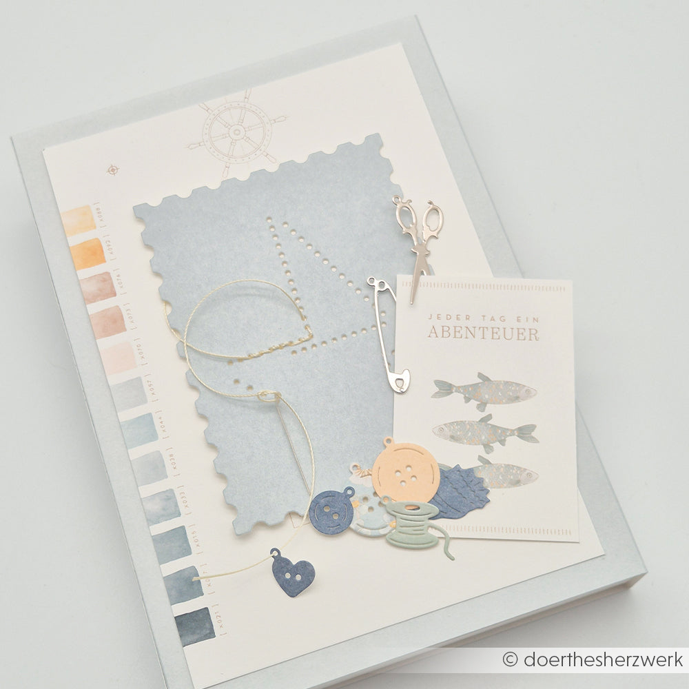 Ein Scrapbook-Layout mit Papieren im maritimen Stil von Alexandra Renke, Garn, Schere, Motivstanzern und Fischillustrationen auf weißem Hintergrund.