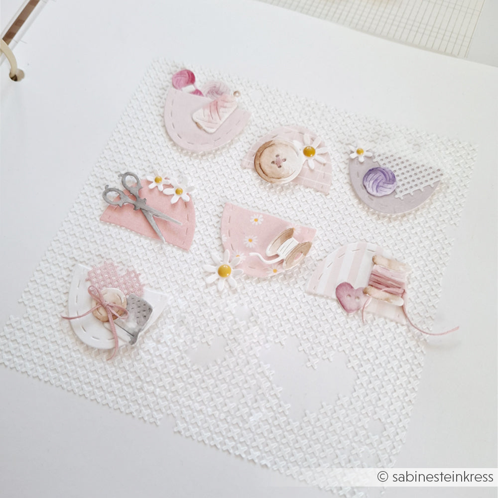 Eine Scrapbook-Seite mit pastellfarbenen Designpapieren von Alexandra Renke zum Thema Nähen, Gänseblümchen, Ausschnitten aus Motivstanzen und einem strukturierten weißen Hintergrund.
