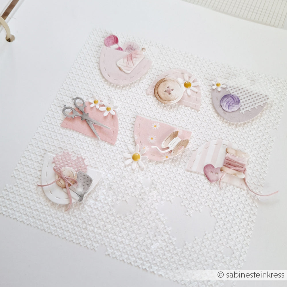 Pastellfarbene Papierausschnitte von Nähutensilien und Blumen, gefertigt aus Designpapieren von Alexandra Renke, sind auf einem strukturierten weißen Hintergrund angeordnet. Die Motivstanze „Stickrahmen Oval“ von Alexandra Renke verleiht der Szene einen charmanten Akzent.
