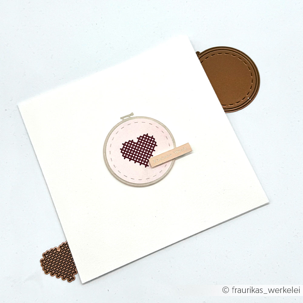 Eine weiße Karte von Alexandra Renke zeigt ein aufgenähtes Herz, das mit der Motivstanze „Stickrahmen Rund“ in einem Stickrahmen eingefasst ist, sowie braune Kreisakzente an zwei Ecken, die aus hochwertigem Designpapier gefertigt sind.