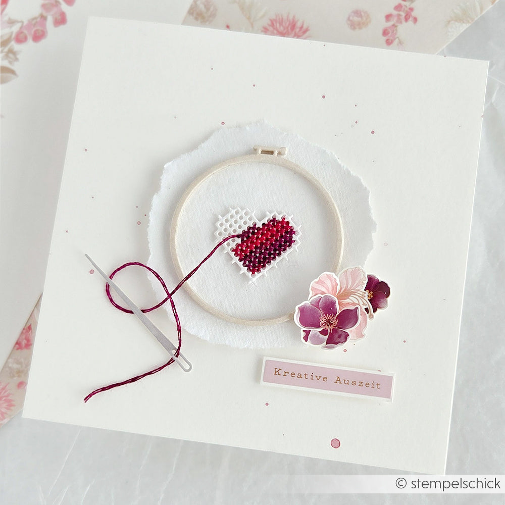 Eine handgefertigte Karte von Alexandra Renke mit Herz-Kreuzstichrahmen, Nadel, rosa Faden, Blumen und Etikett - hergestellt mit Motivstanze Stickrahmen Rund und Stanzsystemen für perfekte Formen.