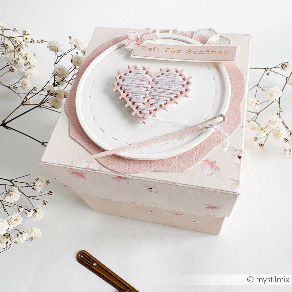 Eine pastellrosa Geschenkbox mit einem aufgestickten Herz und einem deutschen Anhänger, die mit der Motivstanze „Stickrahmen Rund“ von Alexandra Renke gestaltet wurde, ist von zarten weißen Blumen umgeben.